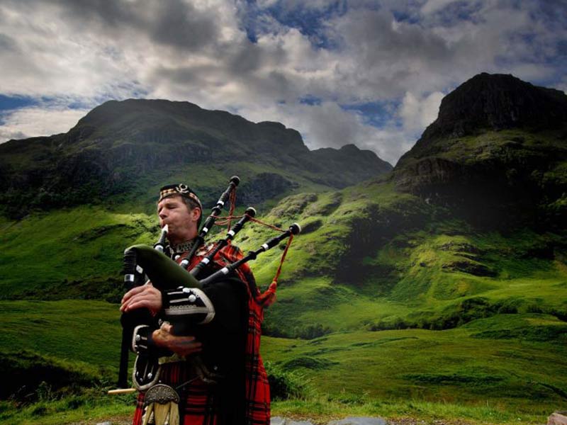 Bagpiper in red tartan kilt playing bagpipes against dramatic mountain landscape.