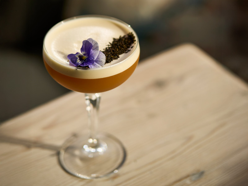An orange foamy cocktail topped with a delicate purple flowers sits on a wooden table in the sun.