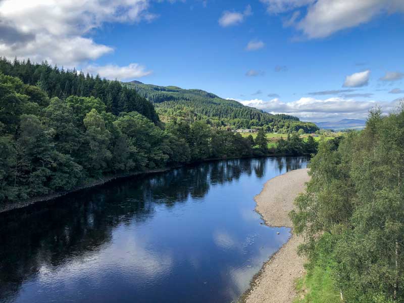 Low aerial photo looking across a quiet, still loch surrounded by lush green trees, taken on a sunny day.