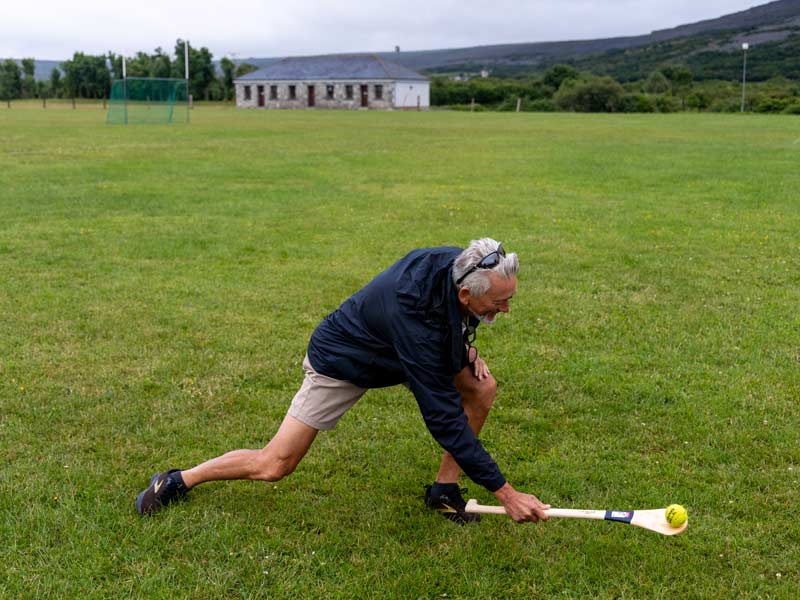 A white man in casual clothing has a go at hurling in a sports field.
