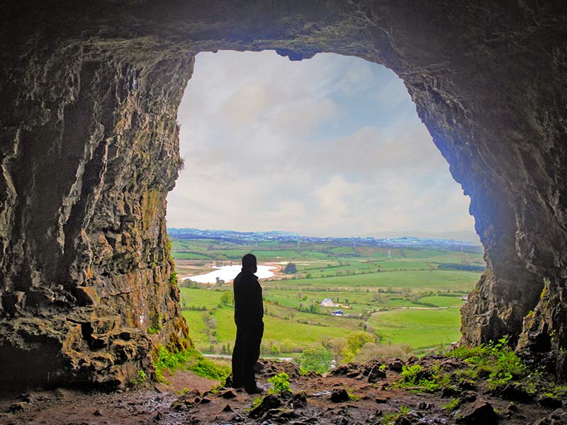 A silhouette of a person stands at the entrance of a rocky cave, overlooking a green countryside with rolling hills