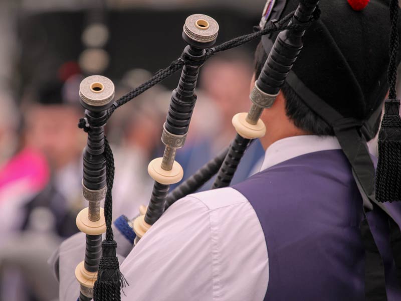 Photo looking over a piper's shoulder to the wooden pipes of the instrument. The background beyond the piper is blurred.