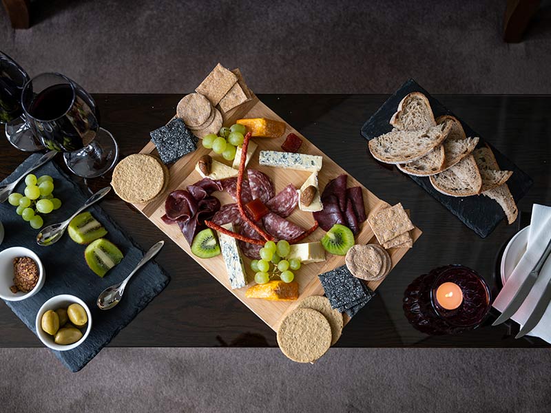 Assorted of meats, cheeses, crackers, grapes, kiwi, olives and wine glasses on a dark wooden surface.
